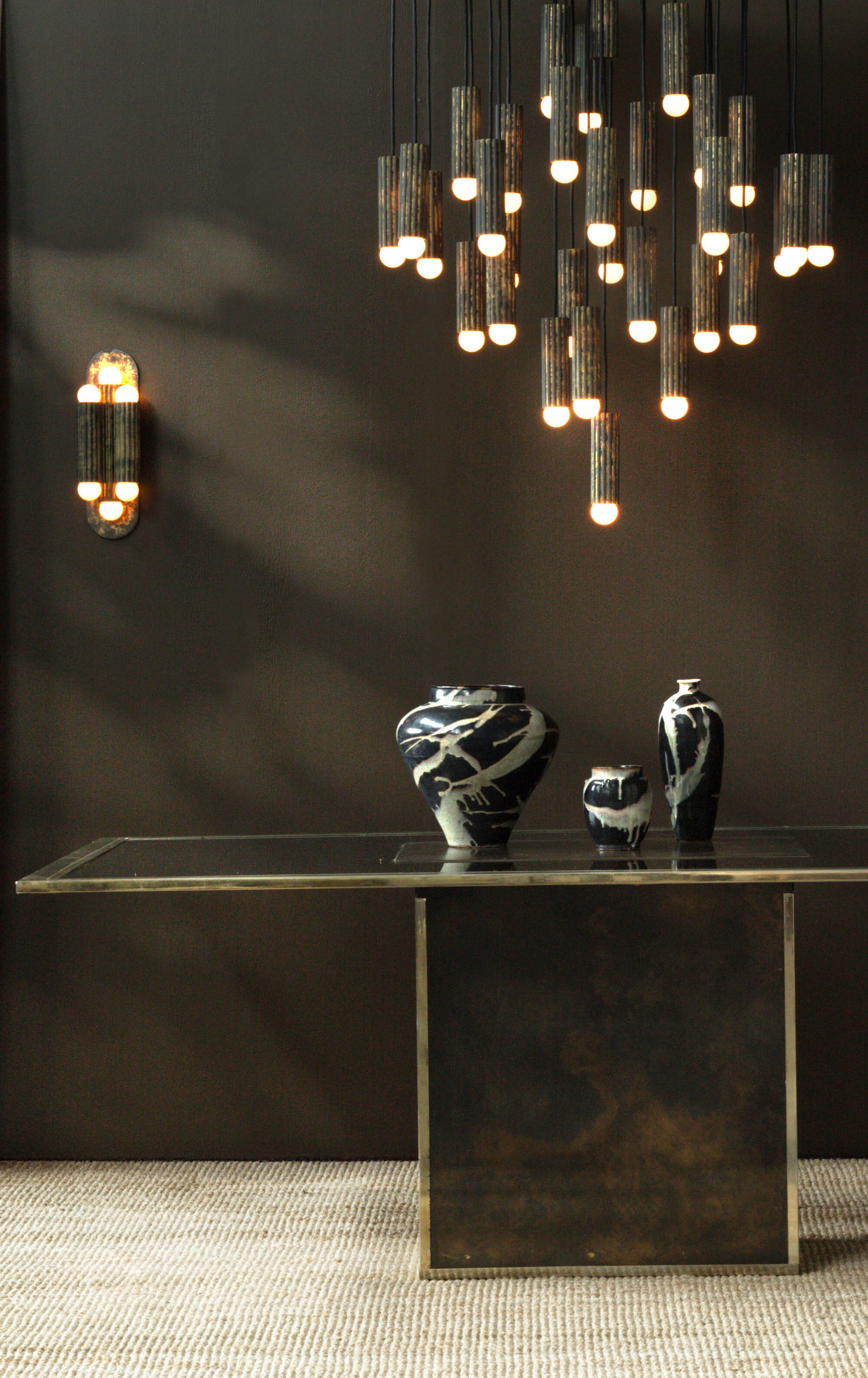 Vesta brass and glass wall sconce with matching chandelier above a sculptural console in a moody luxury interior by Schwung.