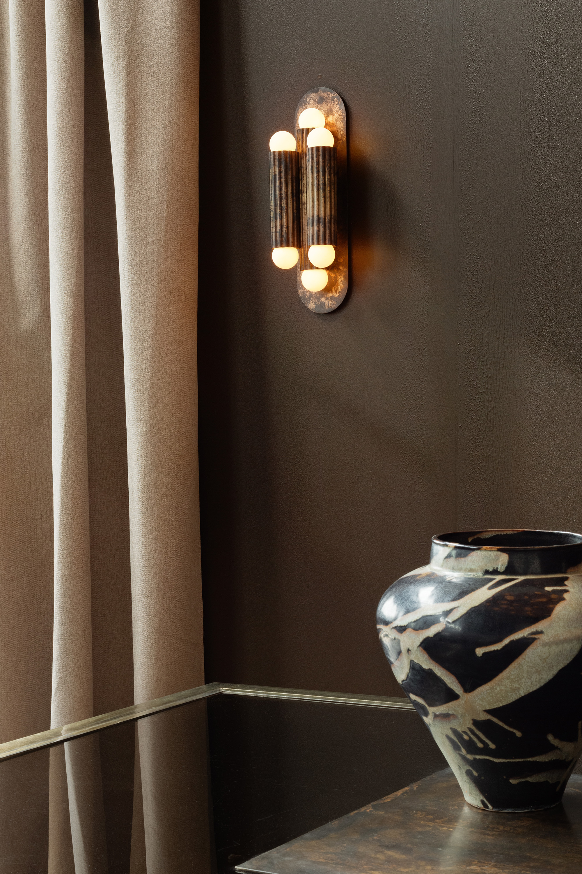 Vesta brass and glass wall sconce in a moody luxury interior with dark walls, soft drapery and decorative vase by Schwung.